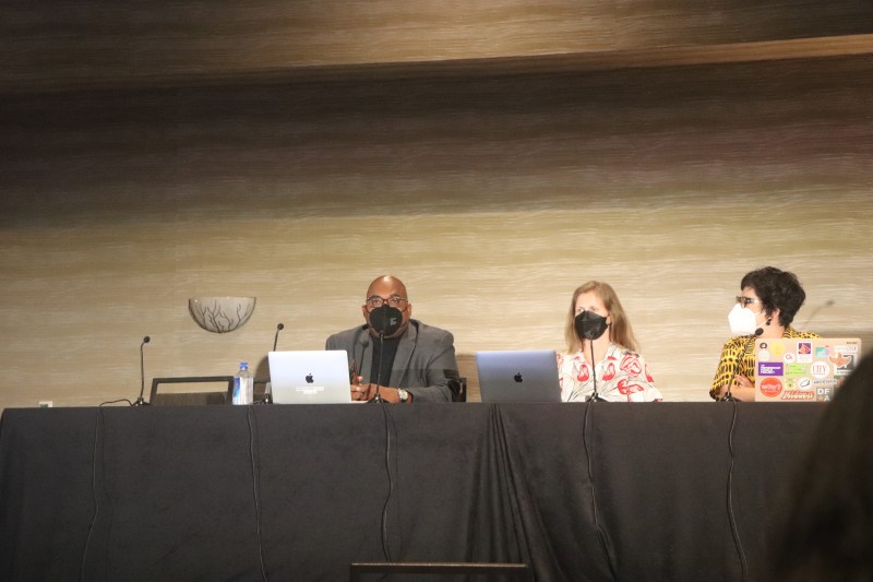 Three panelists sit at a black table with their computers.